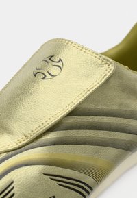 Zlata sintetična natikača z teksturirano površino, črnim črtastim vzorcem in logotipom na zgornji zaponki. Ponaša se z nizkim profilom.