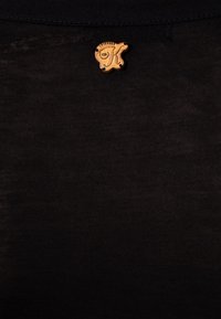 Czarna koszulka z tkaniny o gładkiej teksturze z małą, okrągłą, jasnobrązową metką z logo stylizowanej litery "K" przy dekolcie.