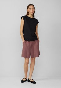 Schwarzes, strukturiertes Kurzarmoberteil kombiniert mit lockeren, hellrosa Shorts. Das Model trägt schwarze Ballerinas. Neutraler Hintergrund.