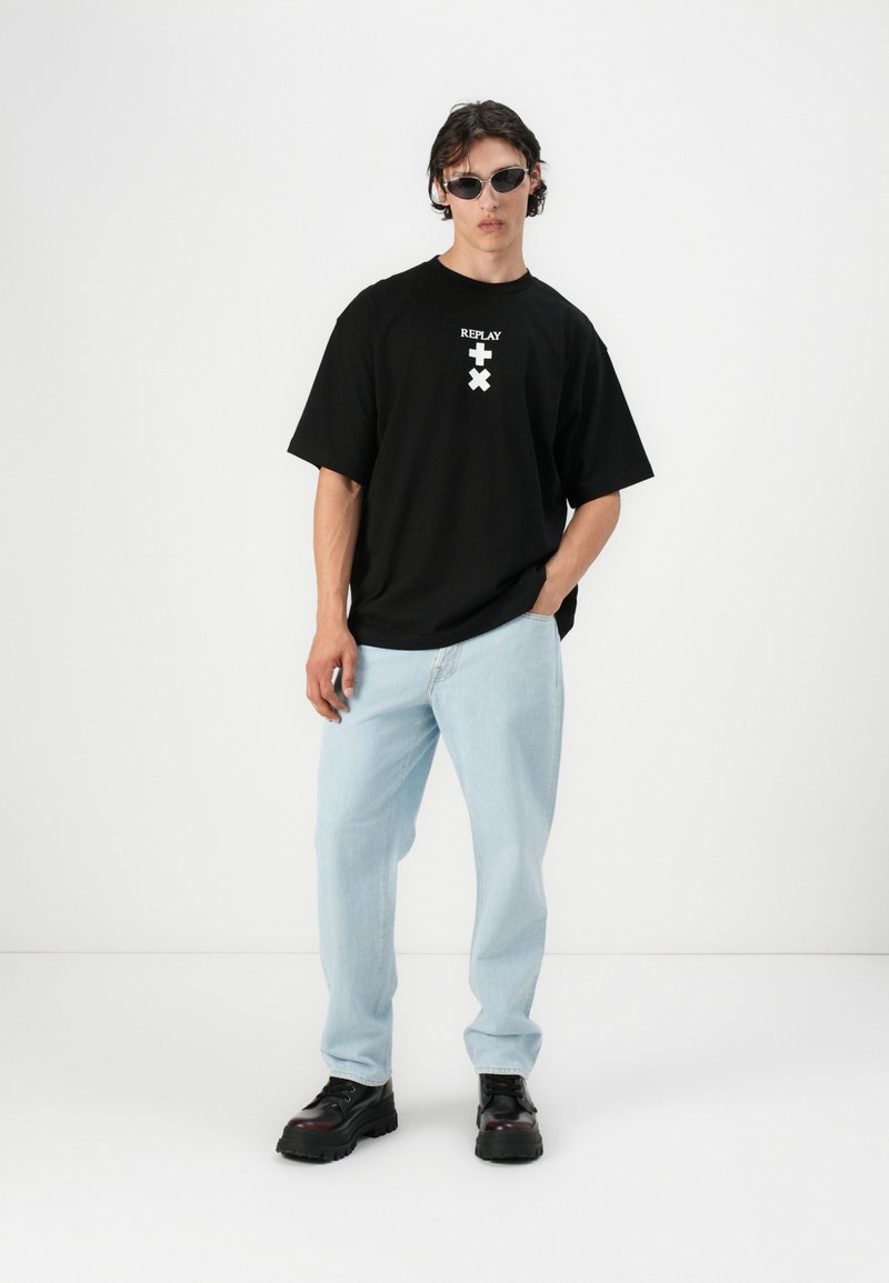 T-shirt noir surdimensionné avec un design de logo blanc, jean en denim bleu clair, et chaussures noires imposantes. Modèle portant de petites lunettes de soleil noires.
