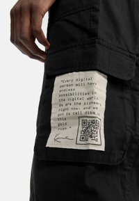 Svart tyg med en texturerad yta, med en vit etikett som har text och en QR-kod fäst vid en ficka på sidan.