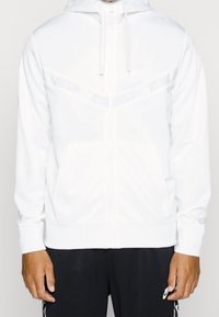 Biała bluza z kapturem zapinana na zamek, wykonana z gładkiego, lekkiego materiału; posiada subtelny wzór kamuflażu na klatce piersiowej oraz przednią kieszeń kangurka.