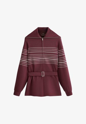 Bordó színű cipzáras dzseki széles gallérral, vízszintes fehér csíkokkal a mellkason és a felső karokon, valamint hozzáillő anyagból készült öv csattal.