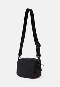 Bolsa transversal preta com formato retangular, textura suave, fecho de zíper e uma alça ajustável com detalhes em logotipo em relevo.