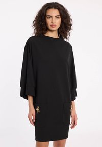 Schwarzes Kleid mit lockerer Passform, Ärmel bis zum Ellbogen, zwei vordere Taschen und goldenen dekorativen Akzenten an jeder Tasche. Glatte Stofftextur.