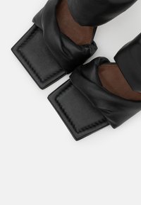 Sandálias de couro pretas com um design superior torcido. Solas quadradas e planas com detalhes castanhos e costura visível ao longo das bordas.