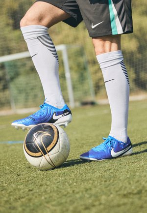 Blauwe Nike voetbalschoenen met een gestructureerde bovenkant, zwarte accenten en witte noppen; een zwart-witte voetbal met gouden details eronder.
