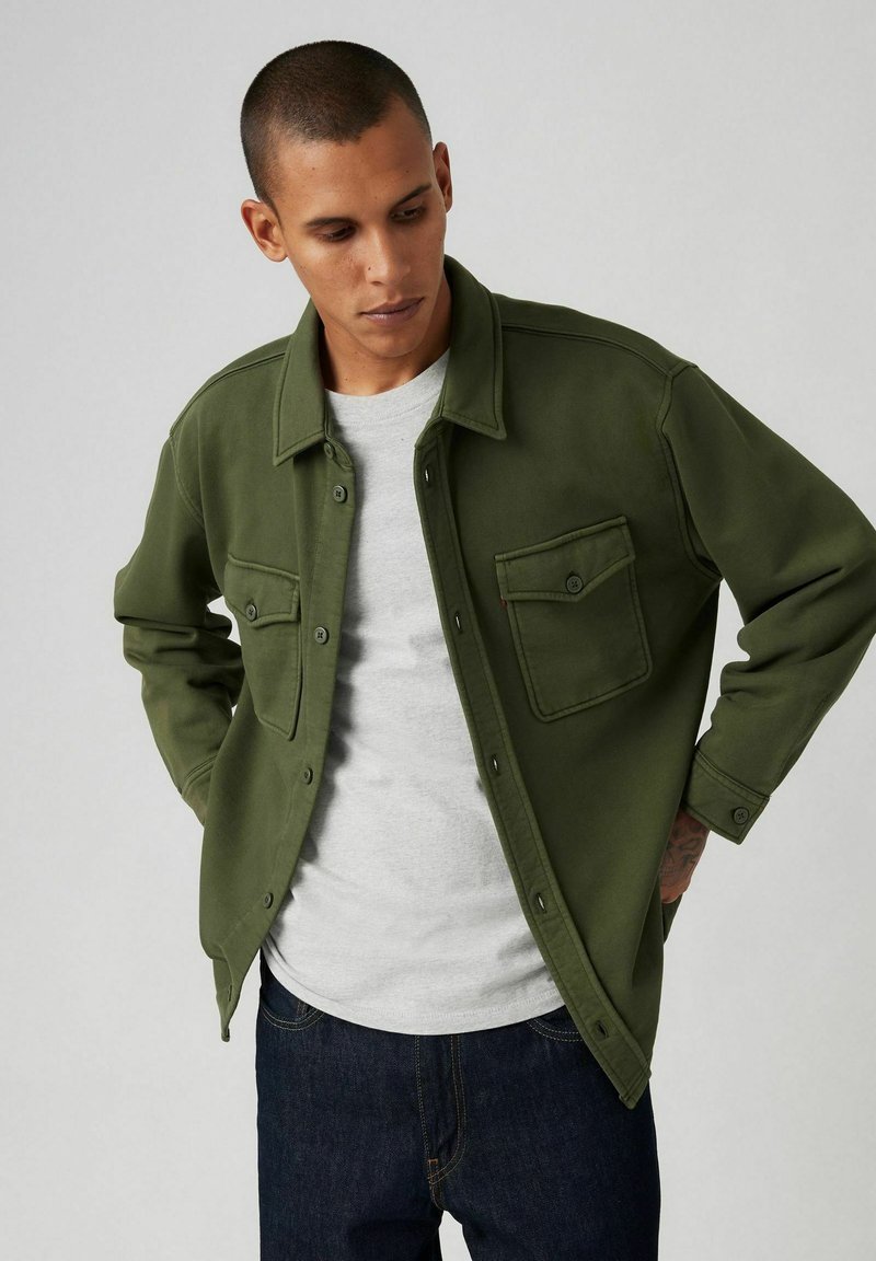 Giacca-camicia verde oliva con bottoni a pressione, due tasche sul petto e vestibilità comoda, indossata sopra una t-shirt grigia e jeans scuri.