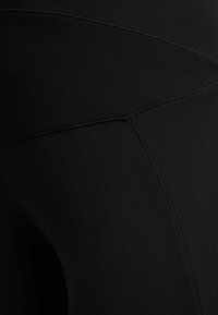 Czarna tkanina leginsów o gładkiej teksturze, dopasowanym kroju i subtelnych detalach szwów wzdłuż boku. Bez widocznych wzorów ani akcentów.