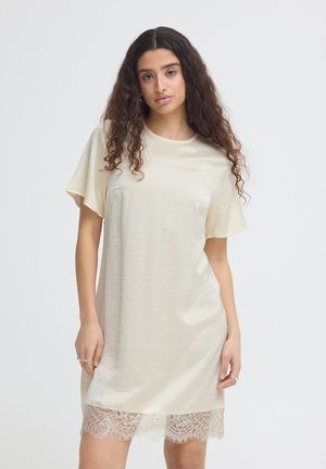 Frau mit langen lockigen Haaren, die ein elfenbeinfarbenes Kurzarmkleid mit Spitzenbesatz am Saum trägt, steht vor einem einfarbigen hellen Hintergrund.