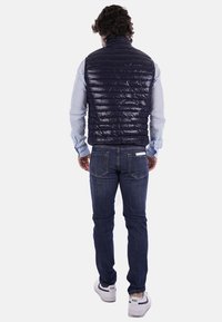 Giubbotto imbottito blu navy con texture trapuntata, indossato sopra una camicia azzurro chiaro, abbinato a jeans scuri e sneakers bianche, visto di spalle.