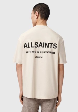 Man with short dark hair wearing a beige ALLSAINTS T-shirt with "SEND ME A POSTCARD" and "LONDON" text on back.
