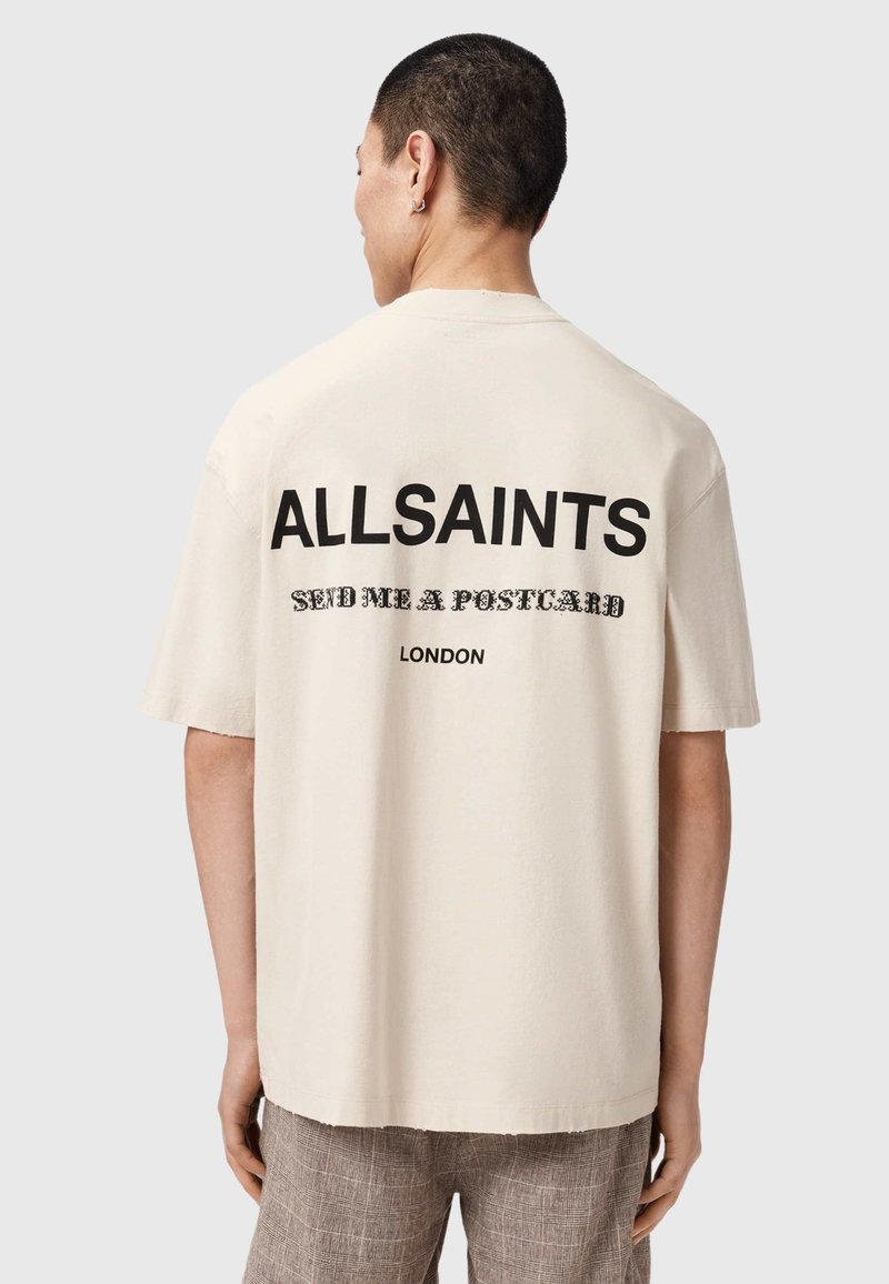 Mann mit kurzen dunklen Haaren, der ein beiges ALLSAINTS T-Shirt trägt, auf dessen Rückseite "SEND ME A POSTCARD" und "LONDON" steht.
