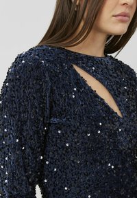Robe en velours bleu marine avec des sequins noirs et argentés denses, présentant un décolleté en trou de serrure et des manches longues. La surface texturée ajoute de la profondeur.