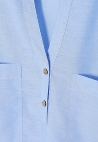 Camicia di lino azzurro chiaro con scollatura a V, due tasche frontali e bottoni di legno marrone. Il tessuto ha una texture liscia e un leggero lucore.