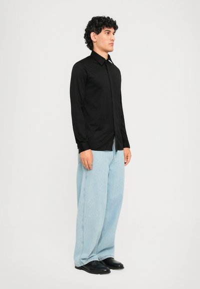 En mann står iført en svart skjorte med lange ermer, lyseblå vide jeans og svarte dressko foran en hvit bakgrunn.