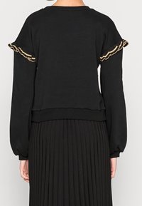 Sudadera negra con mangas con volantes que presenta acentos de hilo dorado. Material suave, corte holgado y cinturilla acanalada, combinada con una falda plisada negra.