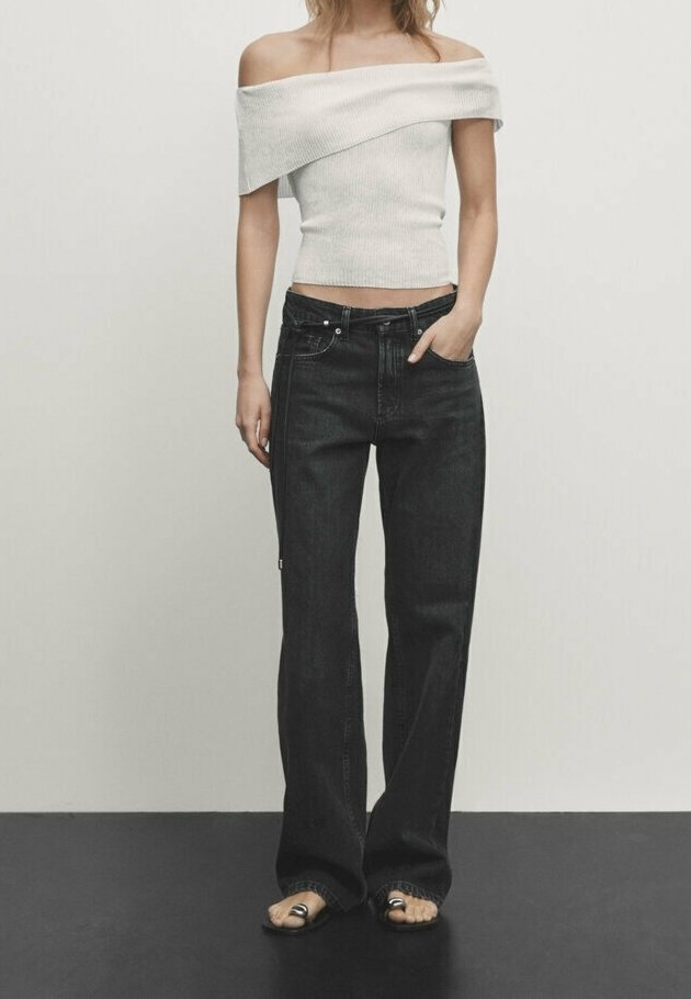 Femme portant un haut blanc à épaules dénudées, un jean noir large taille haute et des sandales noires à bout ouvert, debout devant un fond uni.