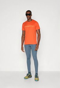 Oranssi DYNAFIT t-paita, kosteutta siirtävää kangasta; harmaat leggingsit; kevyet vihreät ja siniset polkujuoksukengät kuvioiduilla pohjilla; modernit aurinkolasit.