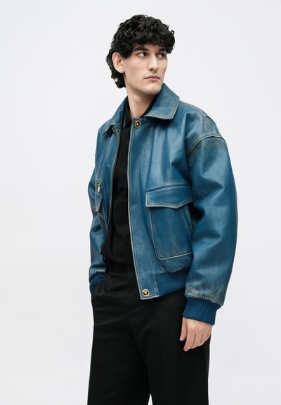 Veste aviateur en cuir bleu avec une fermeture éclair en or, deux poches avant, et des poignets côtelés. Notez la texture légèrement usée et la coupe ajustée.