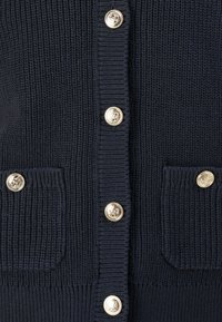 Námořnicky modrý pletený kardigan s žebrovanou texturou, zlatými ozdobnými knoflíky a dvěma předními kapsami s odpovídajícími knoflíkovými detaily.