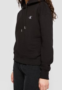 Personne portant un sweat à capuche noir Calvin Klein avec logo sur la poitrine et un jean noir, debout devant un fond uni clair.