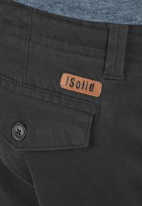 Svarta byxor med en läderlogotypmärkt patch som säger "!Solid." Egenskaper inkluderar knappstängning och strukturerad tyg.