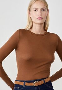 Braunes Langarmshirt aus weichem Material mit einem runden Halsausschnitt, kombiniert mit dunkelblauen Jeans und einem hellbraunen Gürtel.