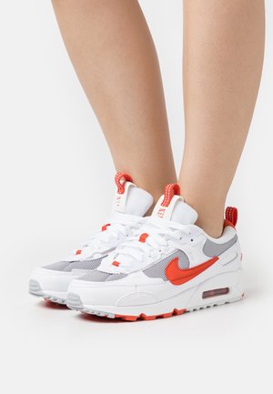 Buty sportowe Nike z białą siateczkową cholewką, szarymi akcentami i czerwonym logo. Posiadają białe sznurowadła oraz widoczną jednostkę powietrzną w podeszwie.