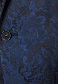 Tissu bleu avec un motif de dentelle florale texturée, doté d'un fermoir à bouton noir. Détails lisses et complexes avec une brillance.