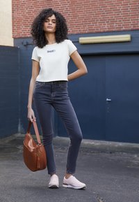 T-shirt beige clair court avec logo ; jean gris taille haute ; chaussures de sport rose pâle ; sac à bandoulière marron avec détail à pampilles.
