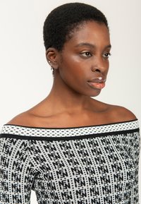 Frau mit kurzen lockigen Haaren, die ein OFF-Shoulder-Musteroberteil trägt, leicht nach rechts gewandt mit einem neutralen Ausdruck vor einem einfarbigen Hintergrund.