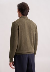 Uomo di spalle che indossa un maglione verde oliva e pantaloni blu scuro su uno sfondo chiaro e semplice.