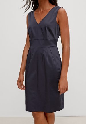 Ærmeløs mørk marineblå V-hals tætsiddende kjole med detaljer ved taljebånd, båret af en stående person mod en ensfarvet baggrund.