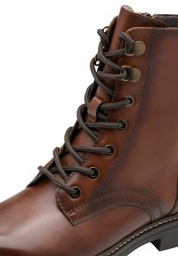 Stivaletto in pelle marrone con punta rotonda, design con lacci, occhielli in metallo e cuciture a contrasto; la superficie testurizzata aggiunge dettagli.