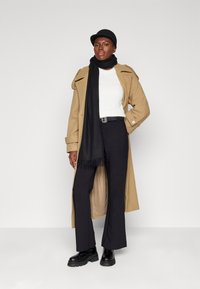 Cappotto beige con spalline, sciarpa nera con frange, maglione bianco, pantaloni neri a vita alta e stivali neri chunky.