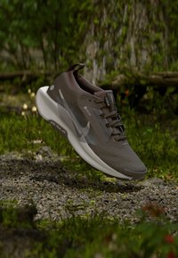 Braun-weißer Nike Trailrunning-Schuh in der Luft über einem Kiesweg in einer grünen Umgebung mit Baumrinde im Hintergrund.