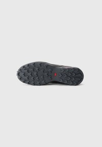 Black hiking shoe sole with deep, chevron-shaped treads and a small red triangle logo in the center on a gray background.