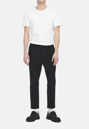 Homme portant un t-shirt blanc uni à manches courtes, un pantalon noir à cordon, des chaussettes blanches et des chaussures noires à lacets, debout devant un fond blanc.