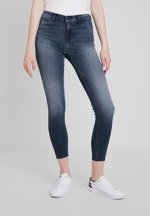 Wąskie, wyblakłe niebieskie dżinsy z postrzępionymi krawędziami, noszone z białymi trampkami i białym topem na jasnoszarym tle.