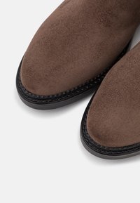 Botines de ante marrón con una textura suave, punta redondeada y suela de goma negra. Presentan costuras decorativas alrededor de la suela.