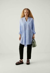 Donna con capelli ricci che indossa una lunga camicia button-up azzurro chiaro, pantaloni neri e sandali neri, mentre tiene in mano una piccola borsa verde.