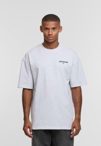 T-shirt oversize grigio chiaro con maniche corte, scollatura rotonda e un dettaglio testuale stampato sul lato sinistro. Tessuto morbido.