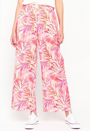 Femme portant un pantalon à jambes larges avec un motif de feuilles tropicales rose, orange et violet et des baskets blanches, debout sur fond blanc.