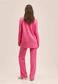 Kobieta z długimi brązowymi włosami stoi tyłem, ubrana w jasnoróżowy garnitur w kropki i białe sandały na obcasie, na jednolitym tle.