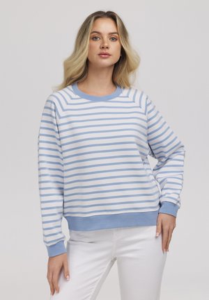 Młoda kobieta z długimi blond włosami, ubrana w białą i jasno-niebieską bluzę w paski oraz białe spodnie, stoi na jednolitym tle.