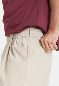 Beige elastic-waist pants with side pockets, worn under a maroon t-shirt, showing a relaxed fit and soft cotton texture.