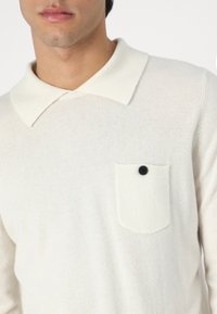 Pulôver de malha em creme claro com um colarinho largo, apresentando um pequeno bolso no peito e detalhes em botão preto. Textura suave, com acabamentos ribados nas bordas.