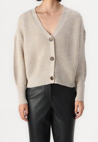 Beige stickad cardigan med V-hals, som har fyra bruna knappar, lös passform och ribbade ärmar. Matchas med svarta läderbyxor.