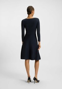 Robe bleu marine à manches longues avec une jupe évasée, en tissu texturé, décolleté rond et taille cintrée. Portée avec des chaussures à talons hauts noires.